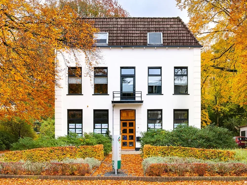Buitenaanzicht van het kantoor van Ditters Makelaars Ede aan de Stationsweg, een karakteristiek wit pand omringd door herfstkleuren en verzorgd groen.
